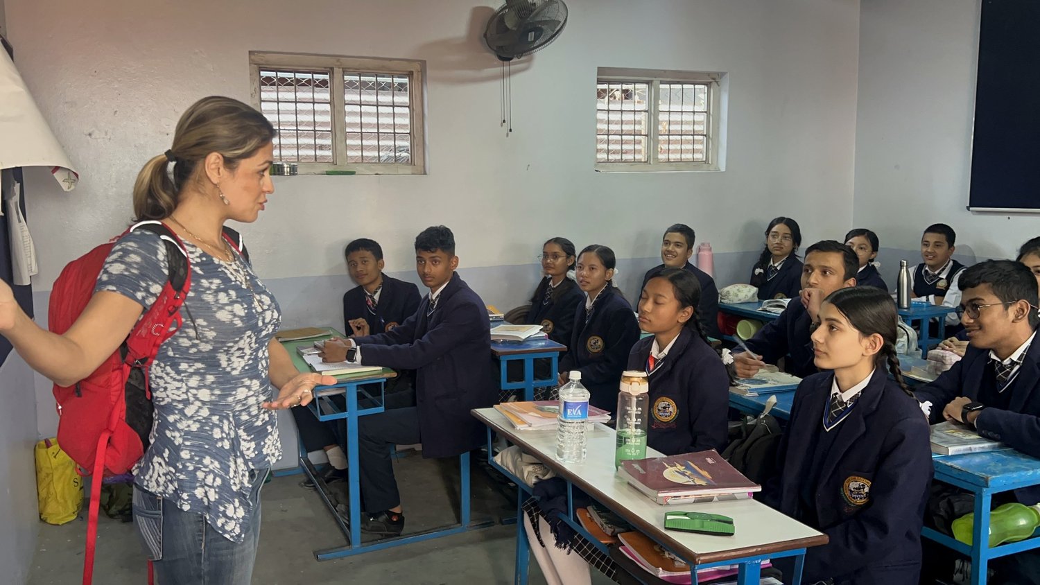Students in school uniform at their desks in a Nepali classroom, a visiting teacher beside them