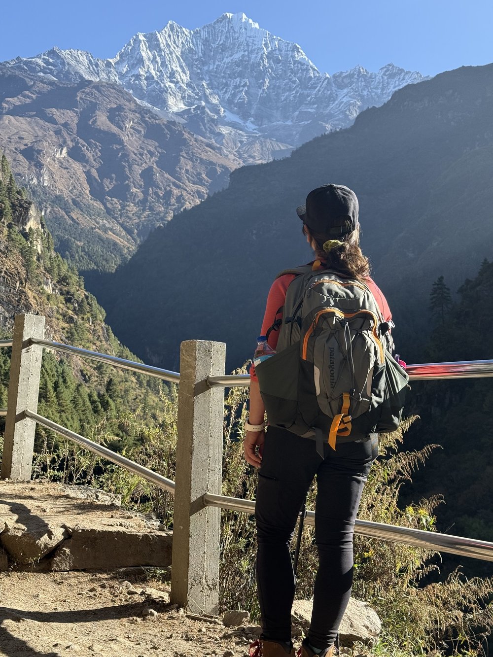 A trekker with a full pack pausing on the trail to look up at a snow-capped Himalayan peak