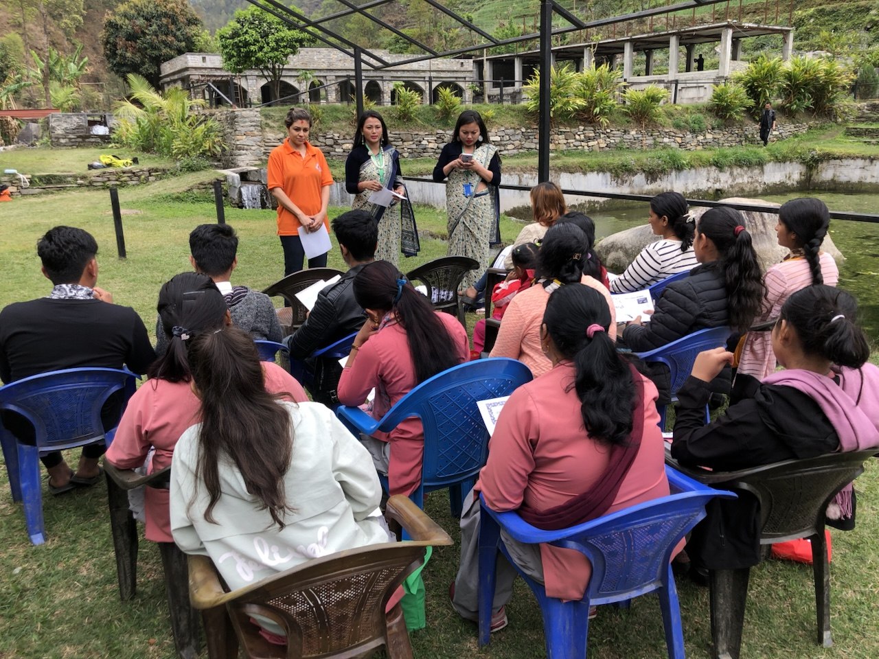 An outdoor workshop on a green hillside: three facilitators standing at the front while a group of young women and men sit in rows of plastic chairs
