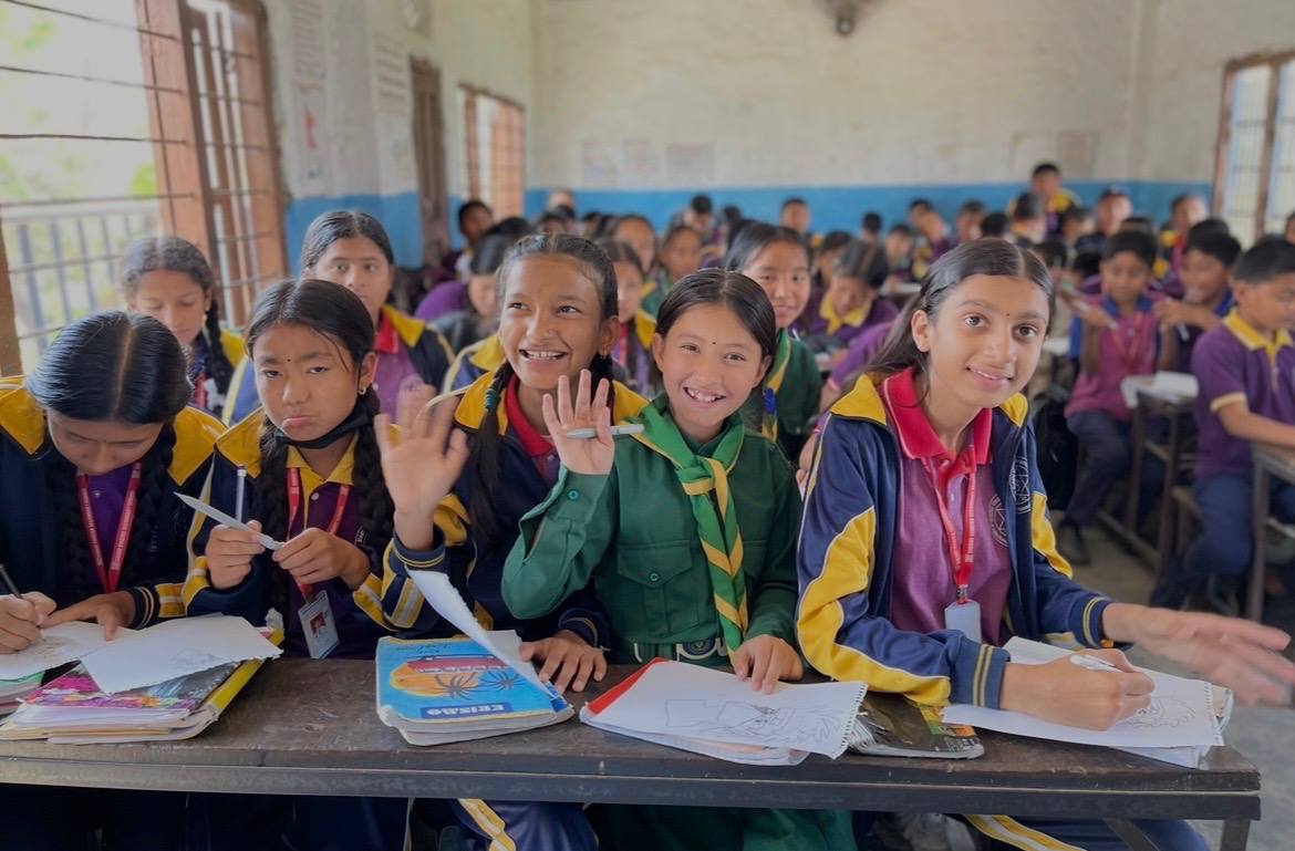 Schoolgirls in uniform at their desks in a Nepali classroom, smiling and waving over their open workbooks