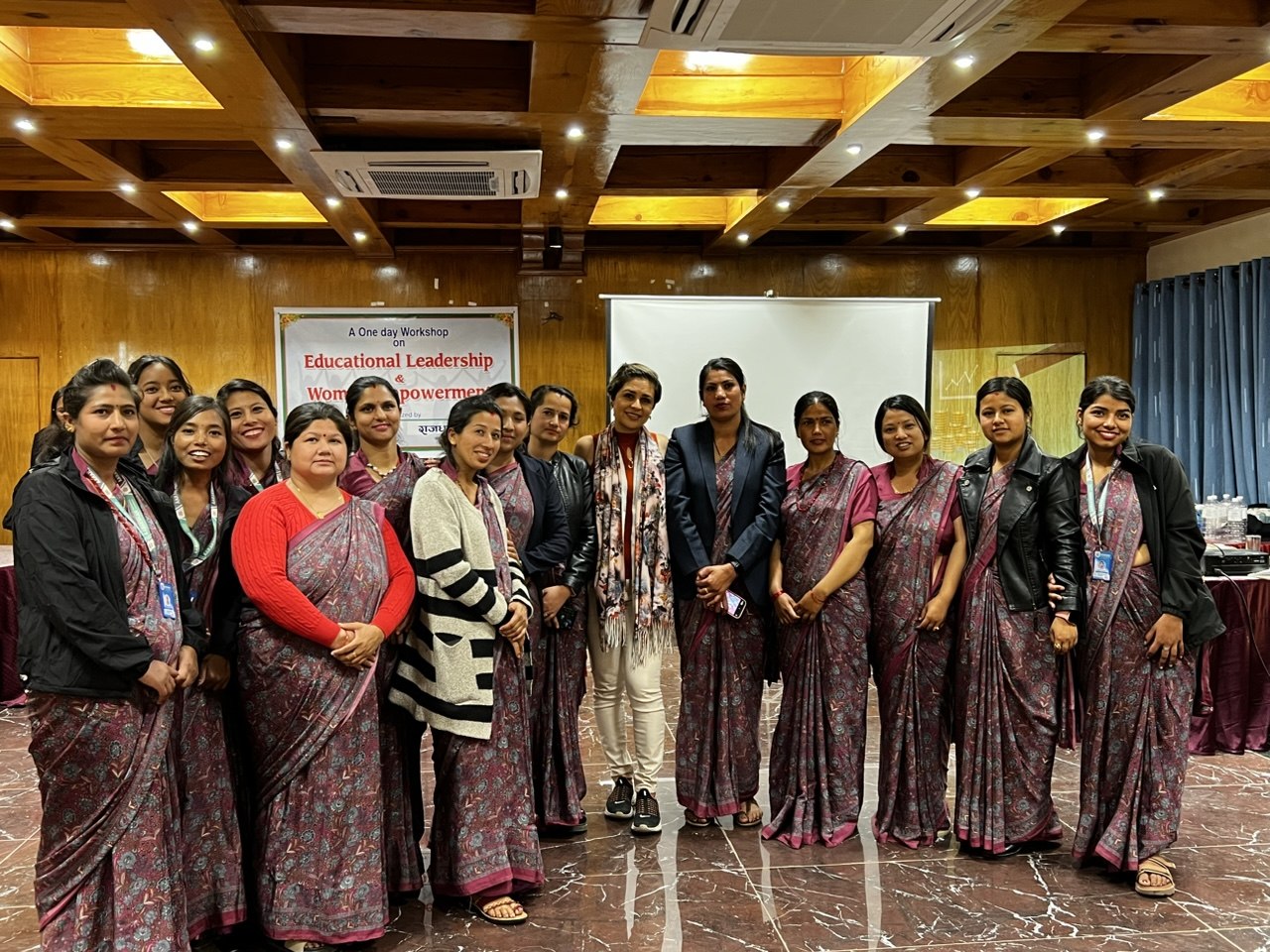 A large group of women, most in matching patterned saris, standing together in front of a banner for a one-day workshop on educational leadership and women's empowerment