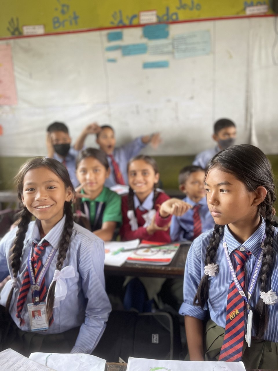 Two schoolgirls in striped uniform and ties at their desks, classmates behind them in a Nepali classroom