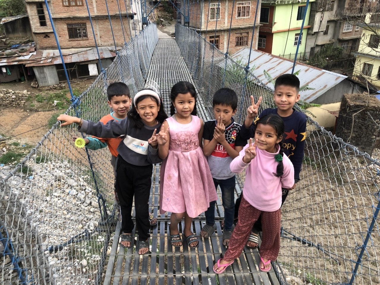 Six children standing together on a suspension footbridge in a Nepali town, smiling and holding up peace signs